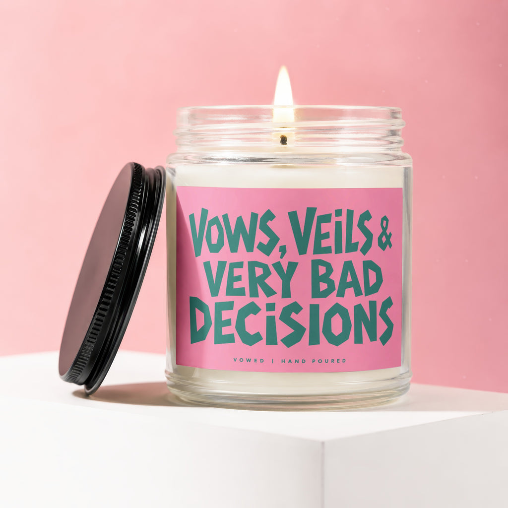 Lit candle in a glass jar with a pink label that reads "VOWS, VEILS & VERY BAD DECISIONS".