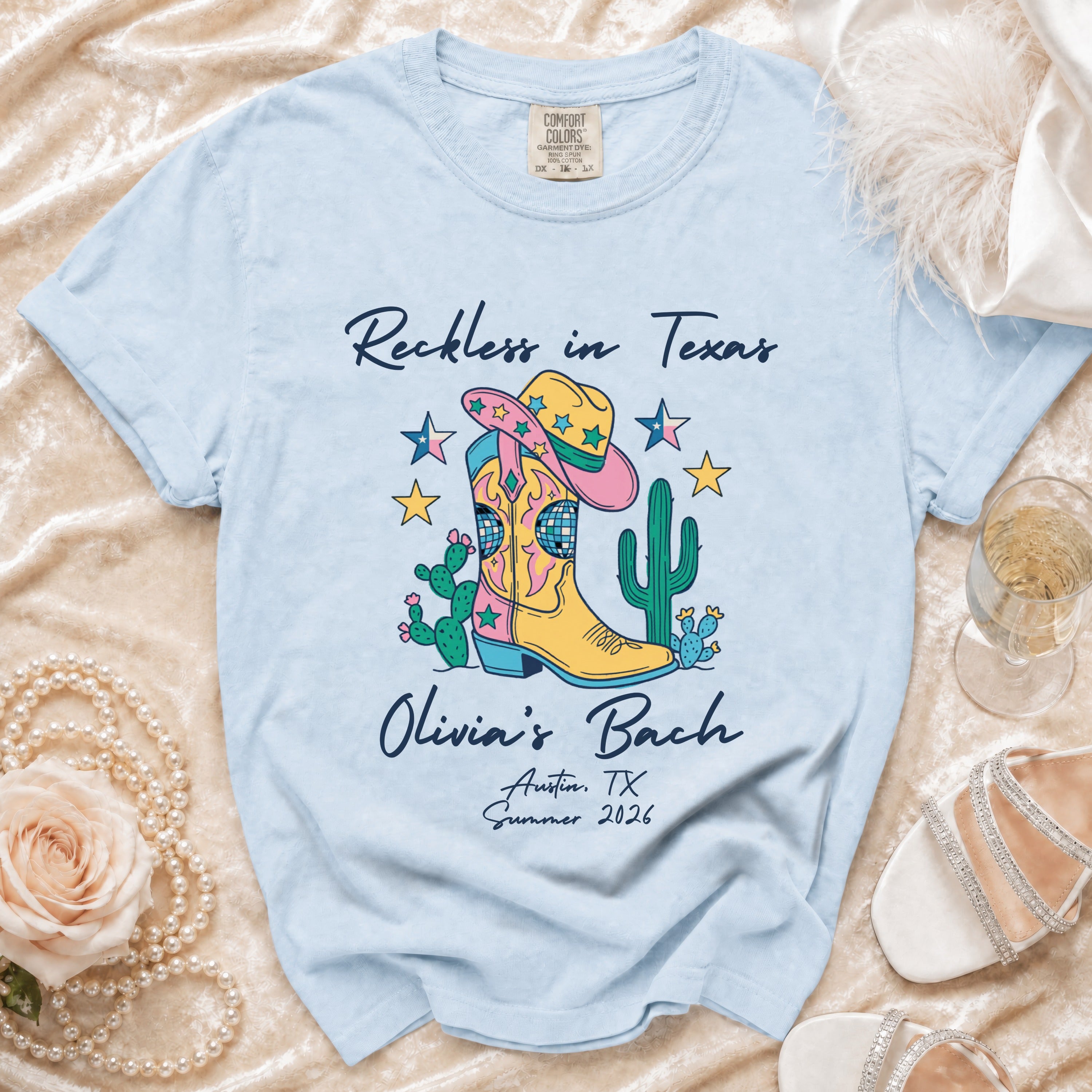 Light blue t-shirt with "Reckless in Texas Olivia's Bach Austin, TX Summer 2026" and boot graphic.