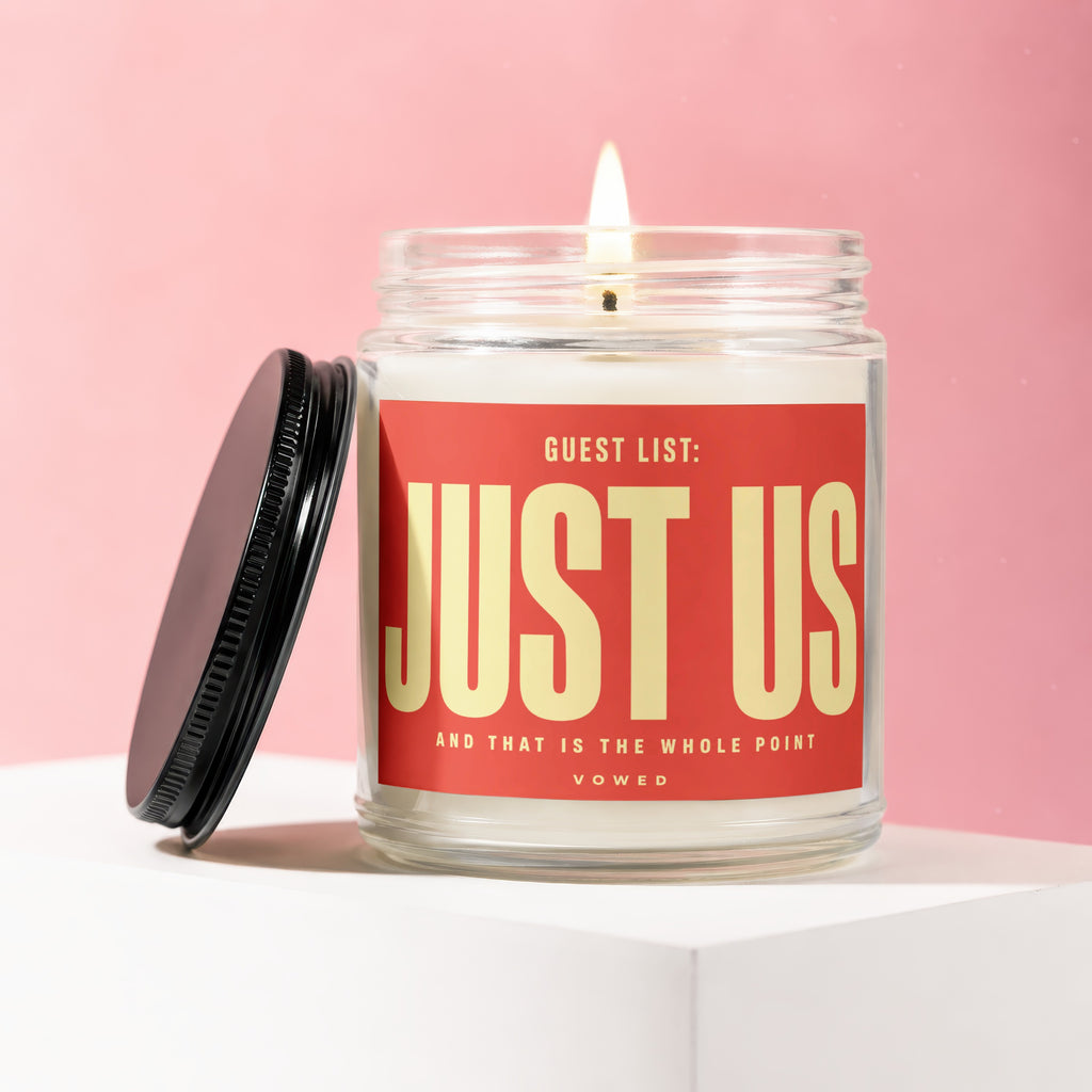 Glass candle with a lit wick and a red label that says "JUST US".
