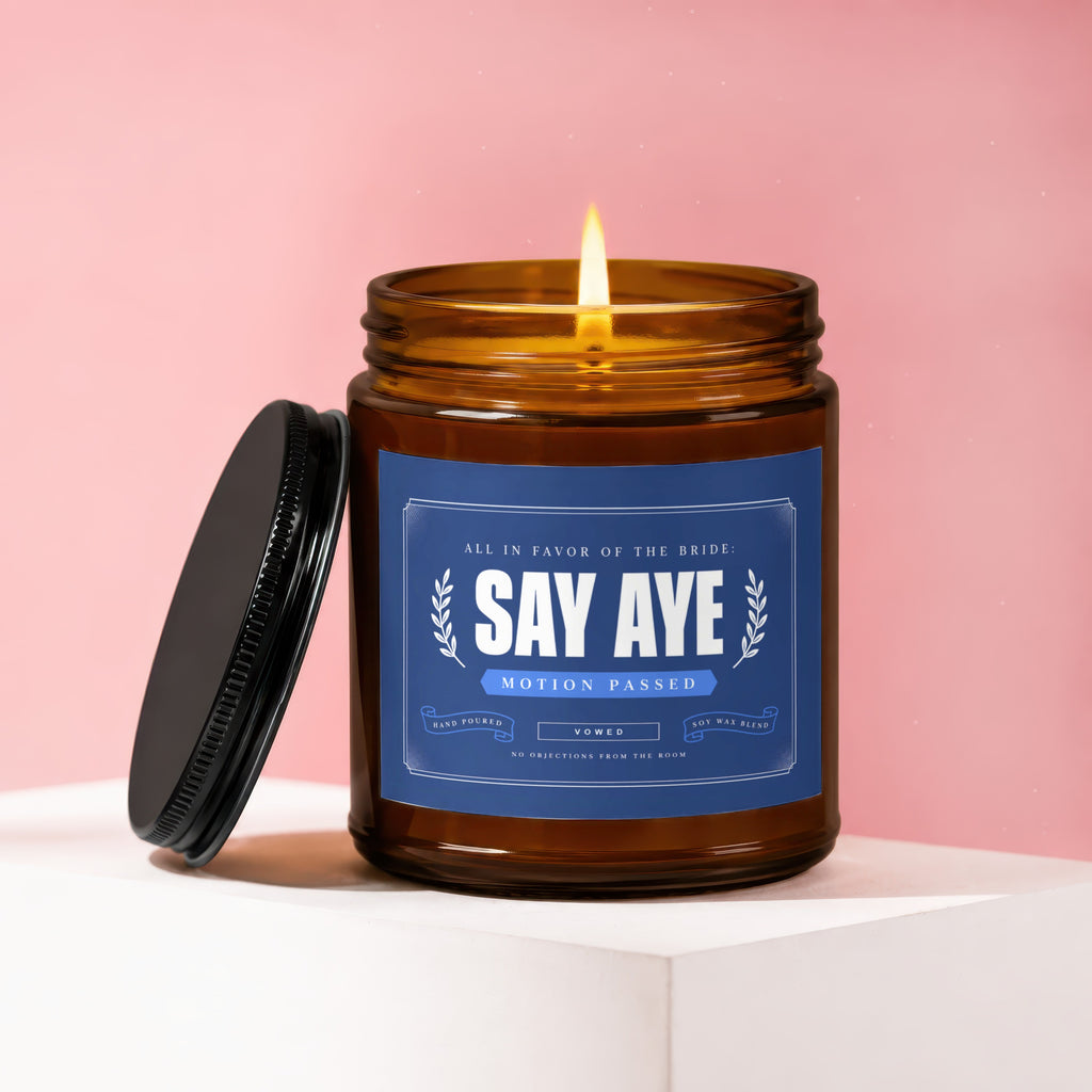Amber jar candle with lit flame, blue label reads "Say Aye Motion Passed Vowed".