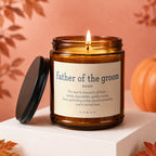 Amber candle with "Father of the Groom" definition on label, lit wick, pumpkin, and fall leaves.