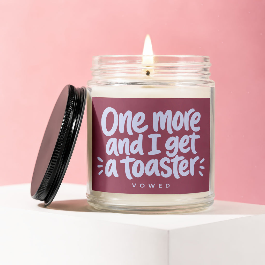 Glass jar candle with lit wick and dark lid, saying "One more and I get a toaster".