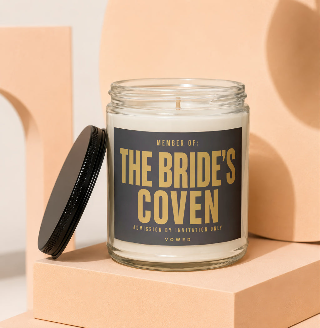 Glass candle with "The Bride's Coven" text and lid rest on stacked blocks.