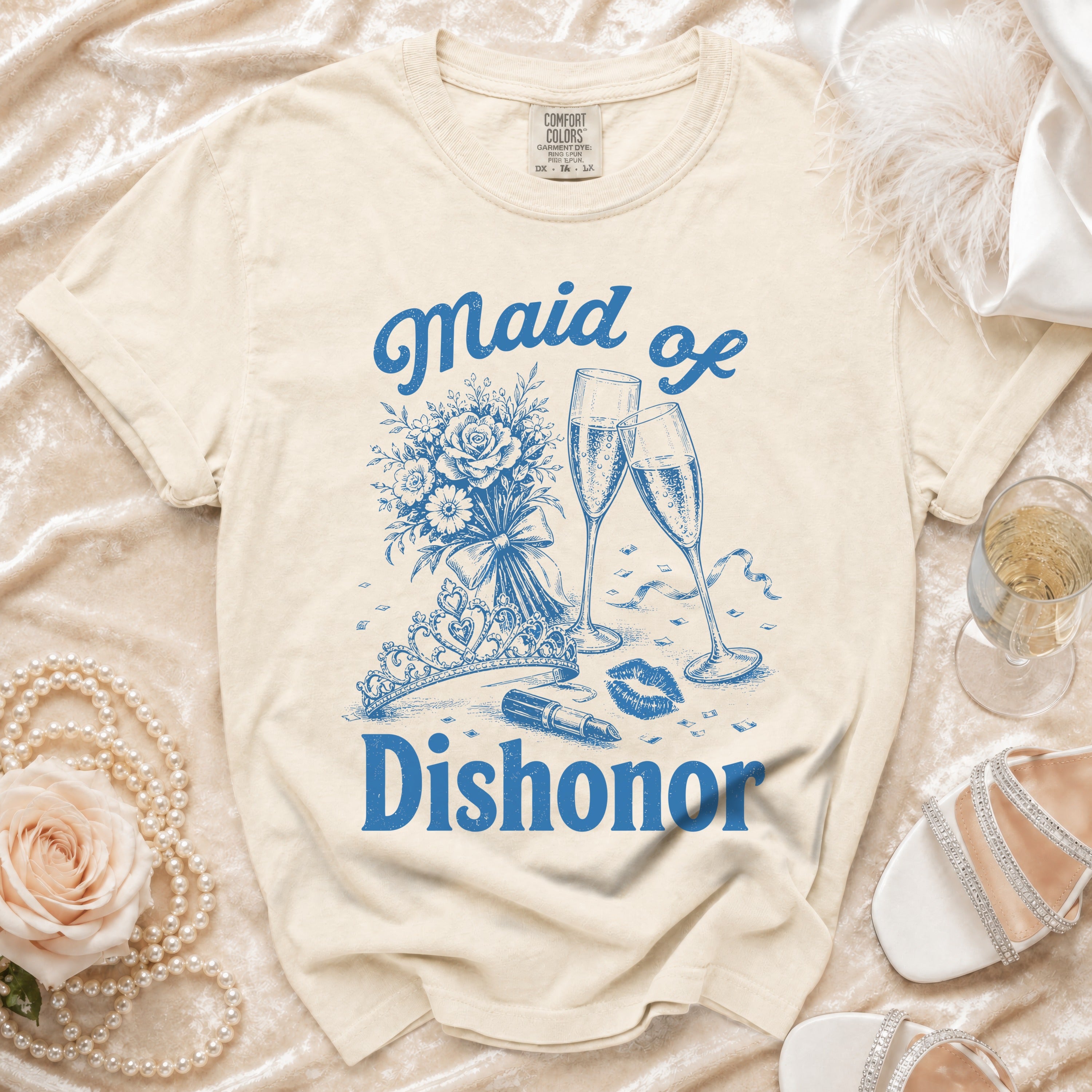 Cream t-shirt with blue "Maid of Dishonor" text, bouquet, tiara, lipstick, and champagne flutes.