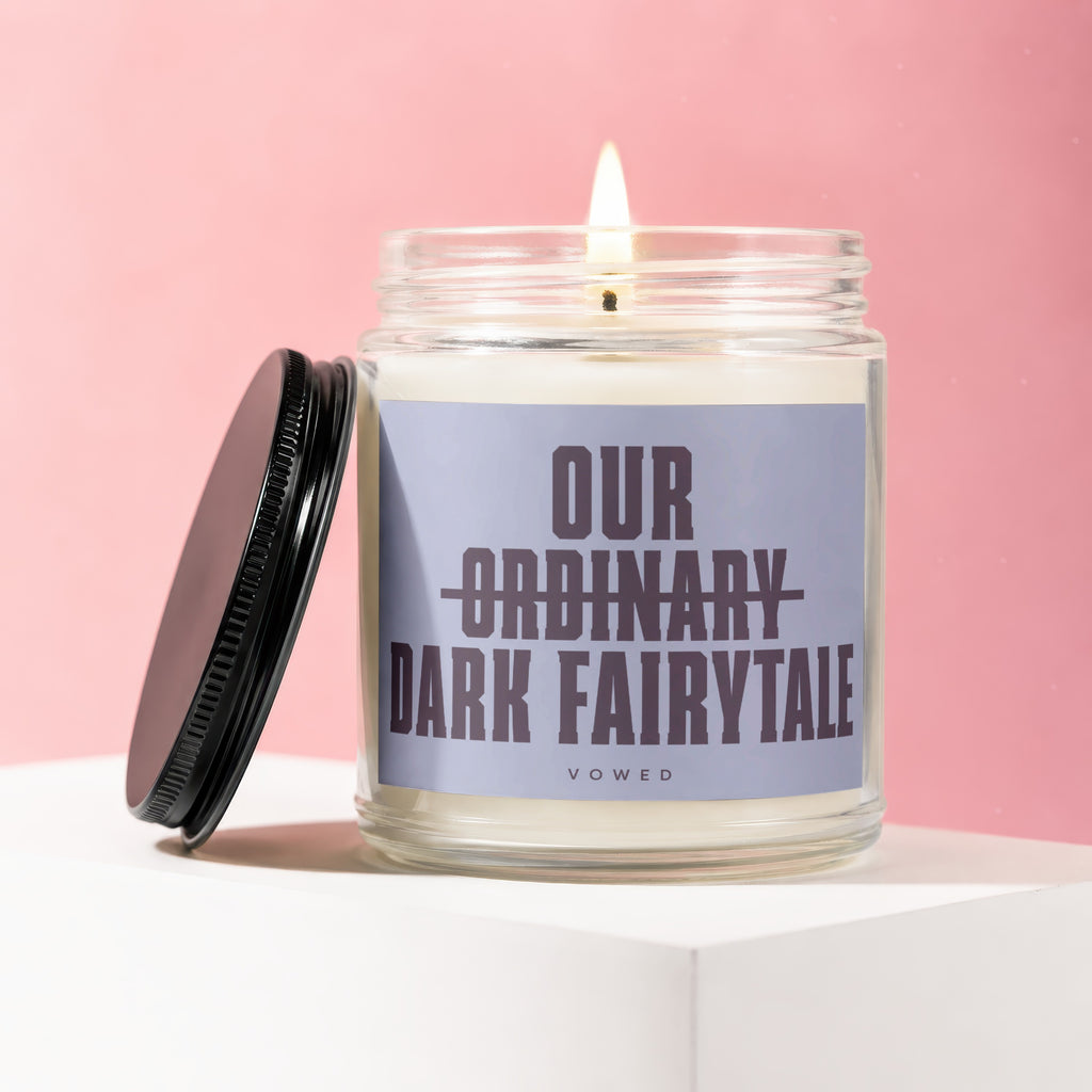 Glass candle with lit wick, lid to the left, label reads "Our Ordinary Dark Fairytale Vowed".