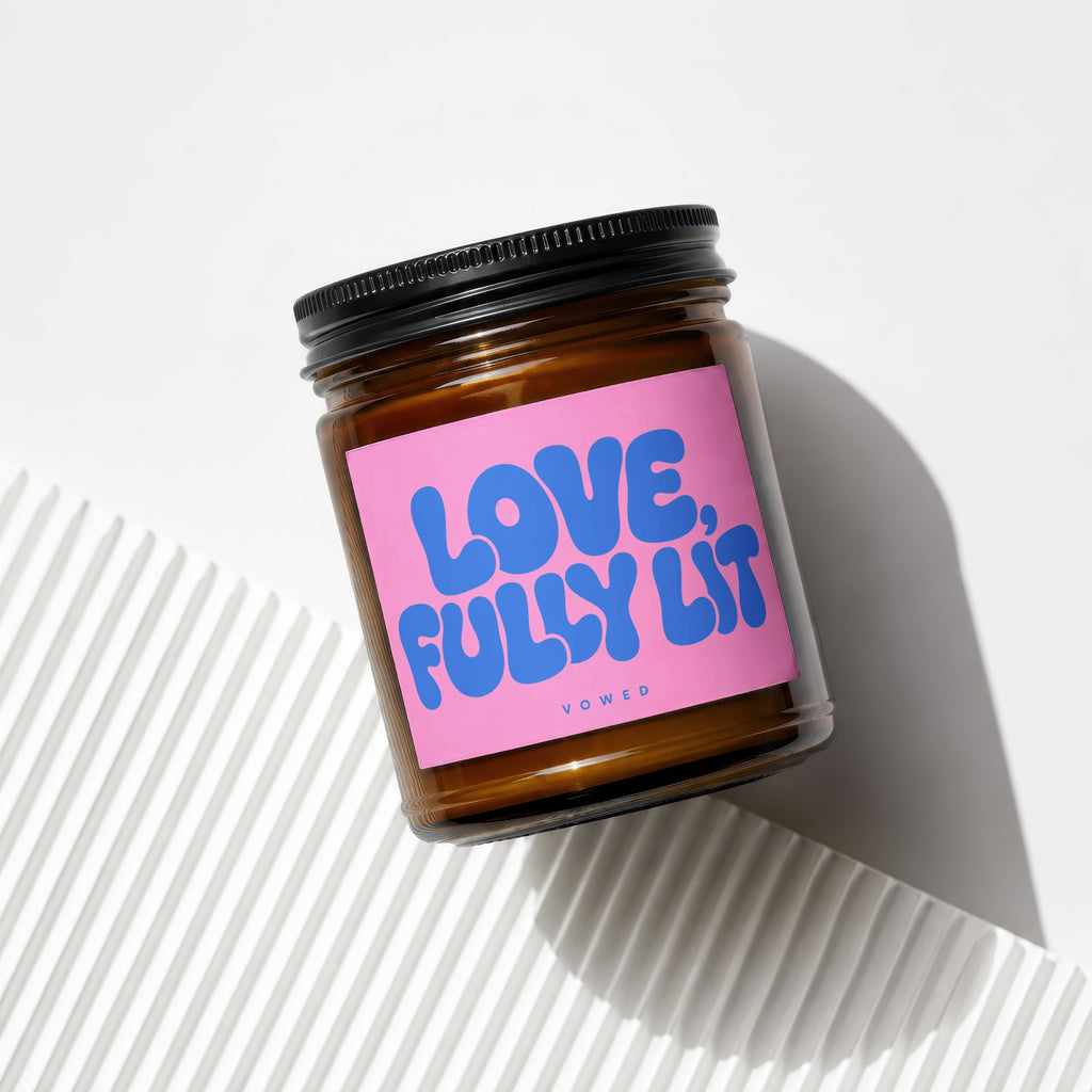 Glass candle jar with black lid, featuring a pink label with blue text that reads "LOVE, FULLY LIT".