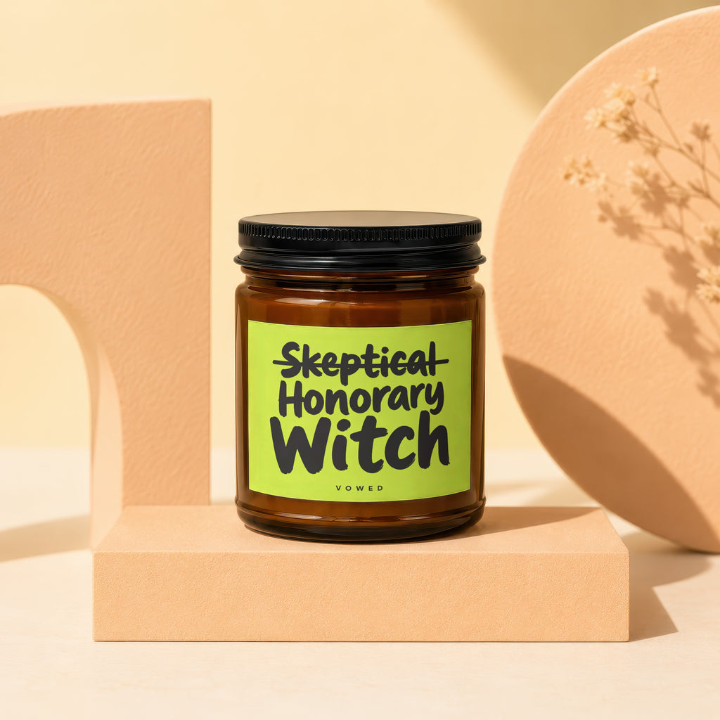 Amber glass candle with black lid, "Skeptical Honorary Witch VOWED" on a lime green label.