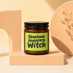 Amber glass candle with black lid, "Skeptical Honorary Witch VOWED" on a lime green label.
