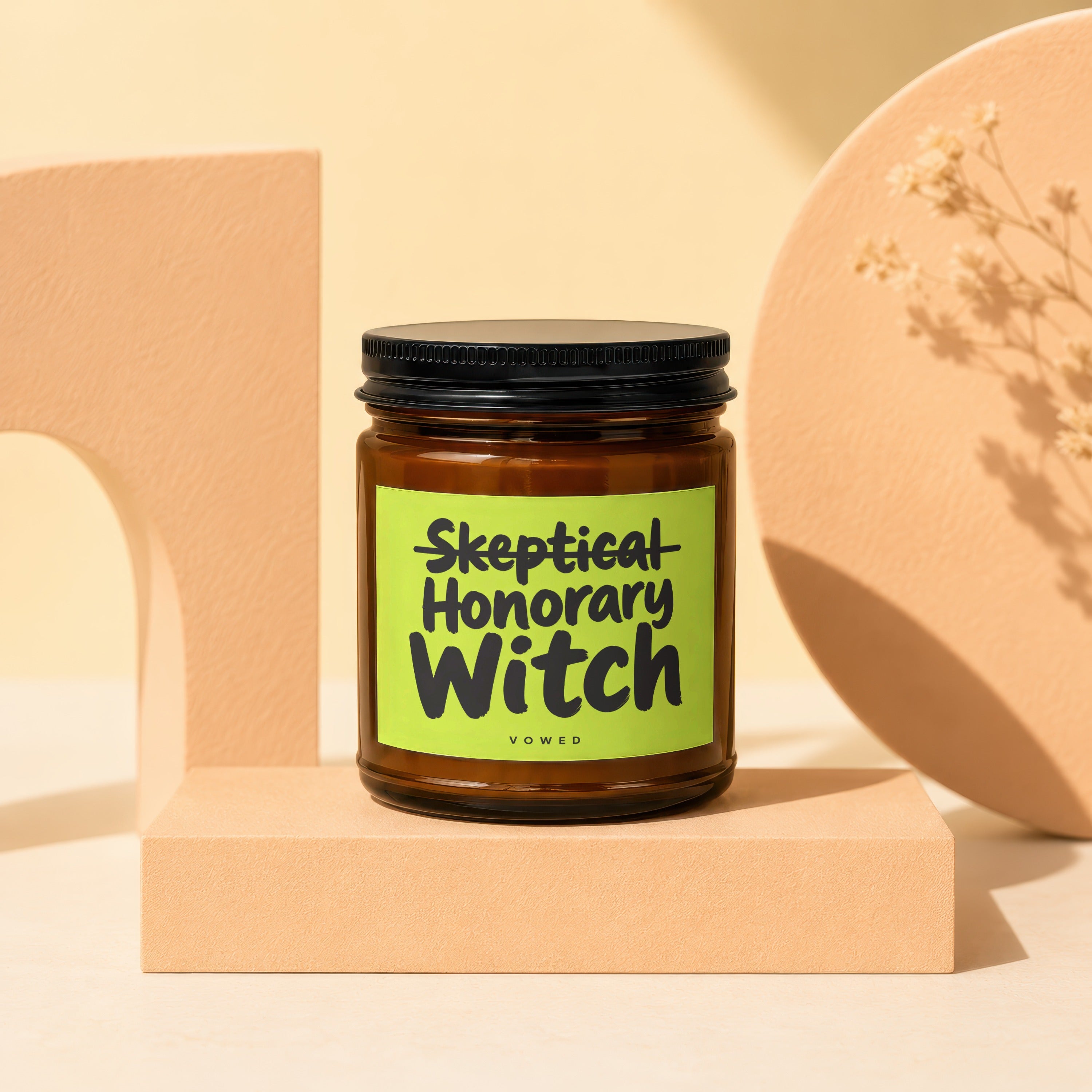 Amber glass candle with black lid, "Skeptical Honorary Witch VOWED" on a lime green label.