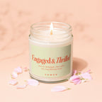 Glass jar candle with lit wick, label reads "Engaged & Thrilled" and "VOWED".