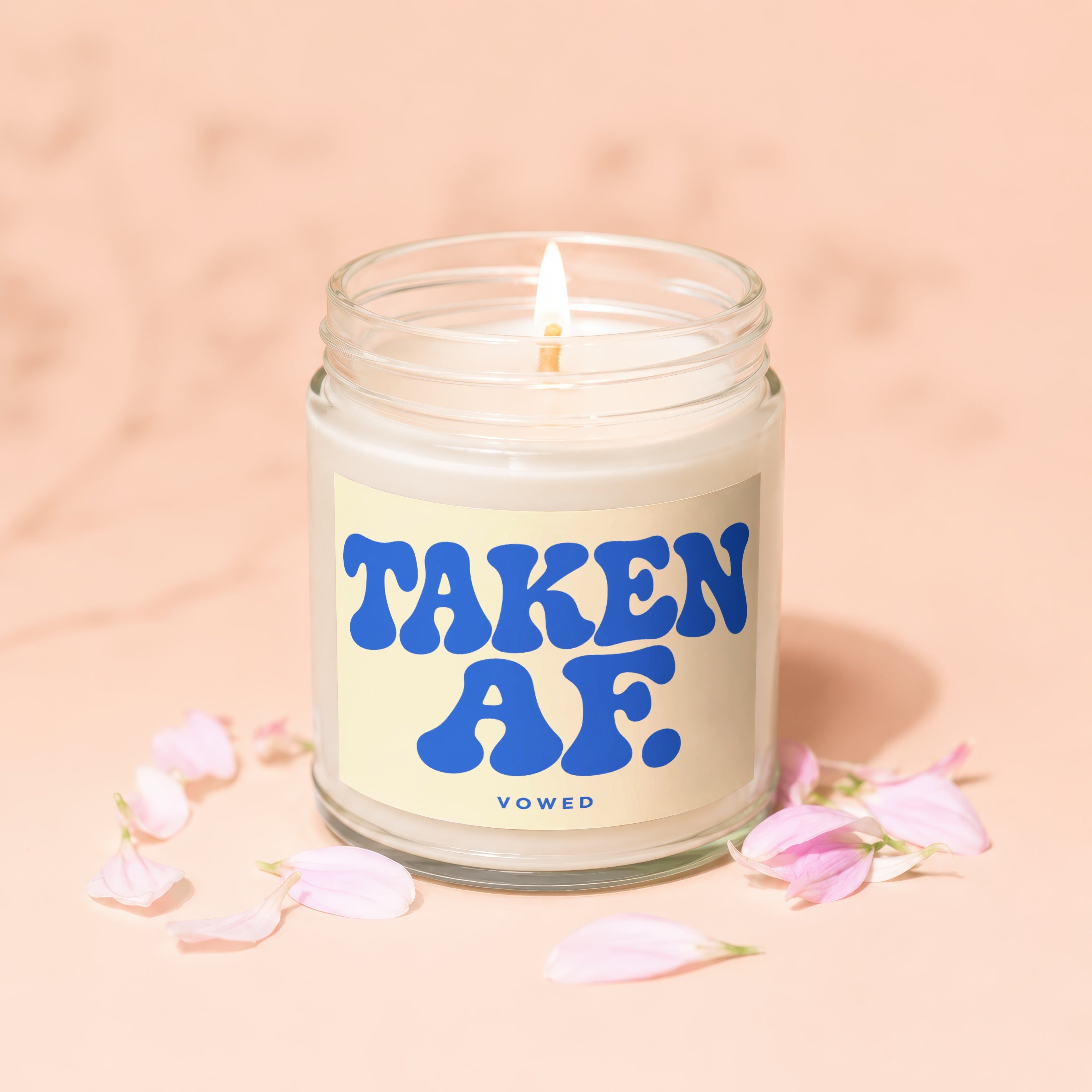 Glass jar candle with blue text "TAKEN AF. VOWED" surrounded by pink petals.