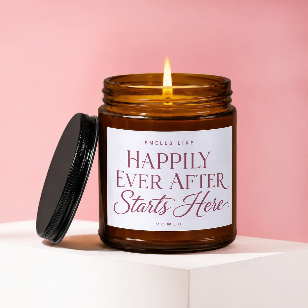 Brown glass candle with lit wick, lid off, reading "Smells Like Happily Ever After Starts Here".