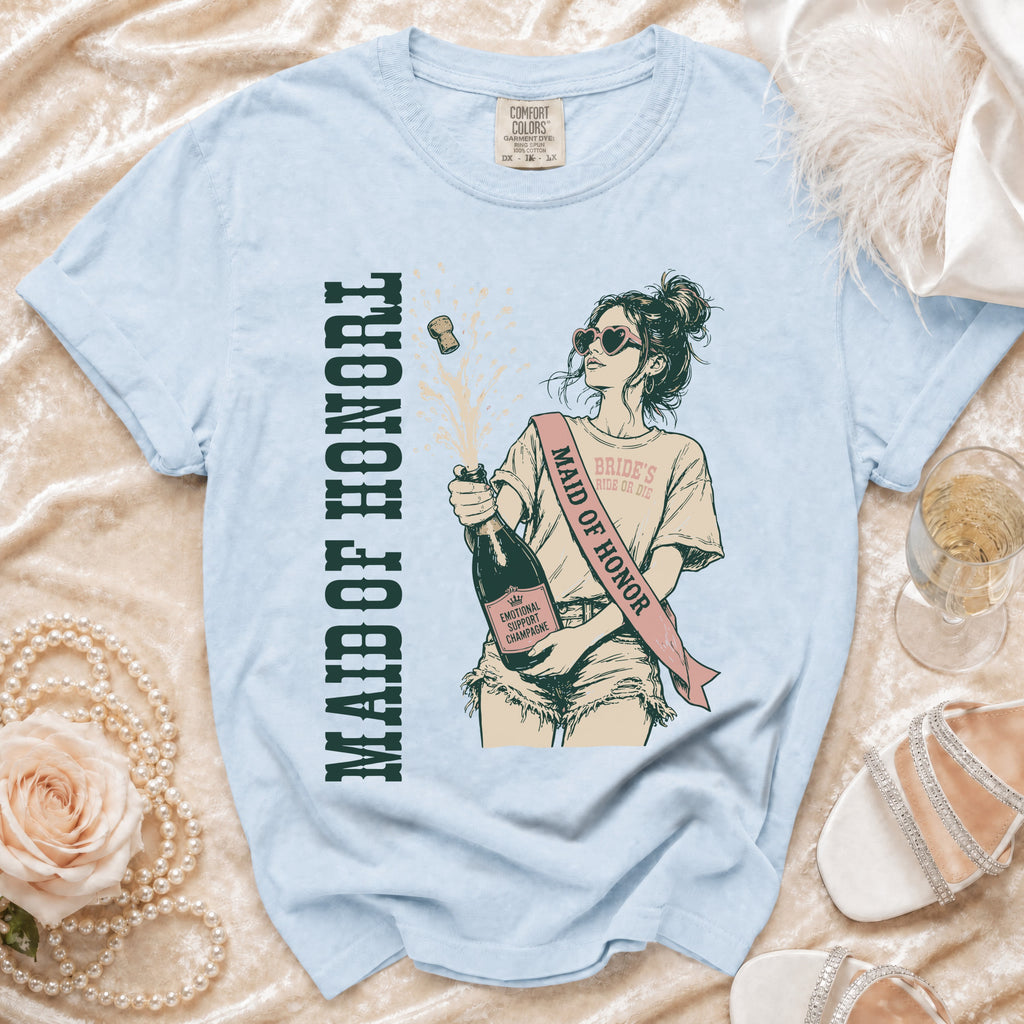 Light blue t-shirt with illustration of a woman popping champagne. Text reads "MAID OF HONOR" and "BRIDE'S RIDE OR DIE".