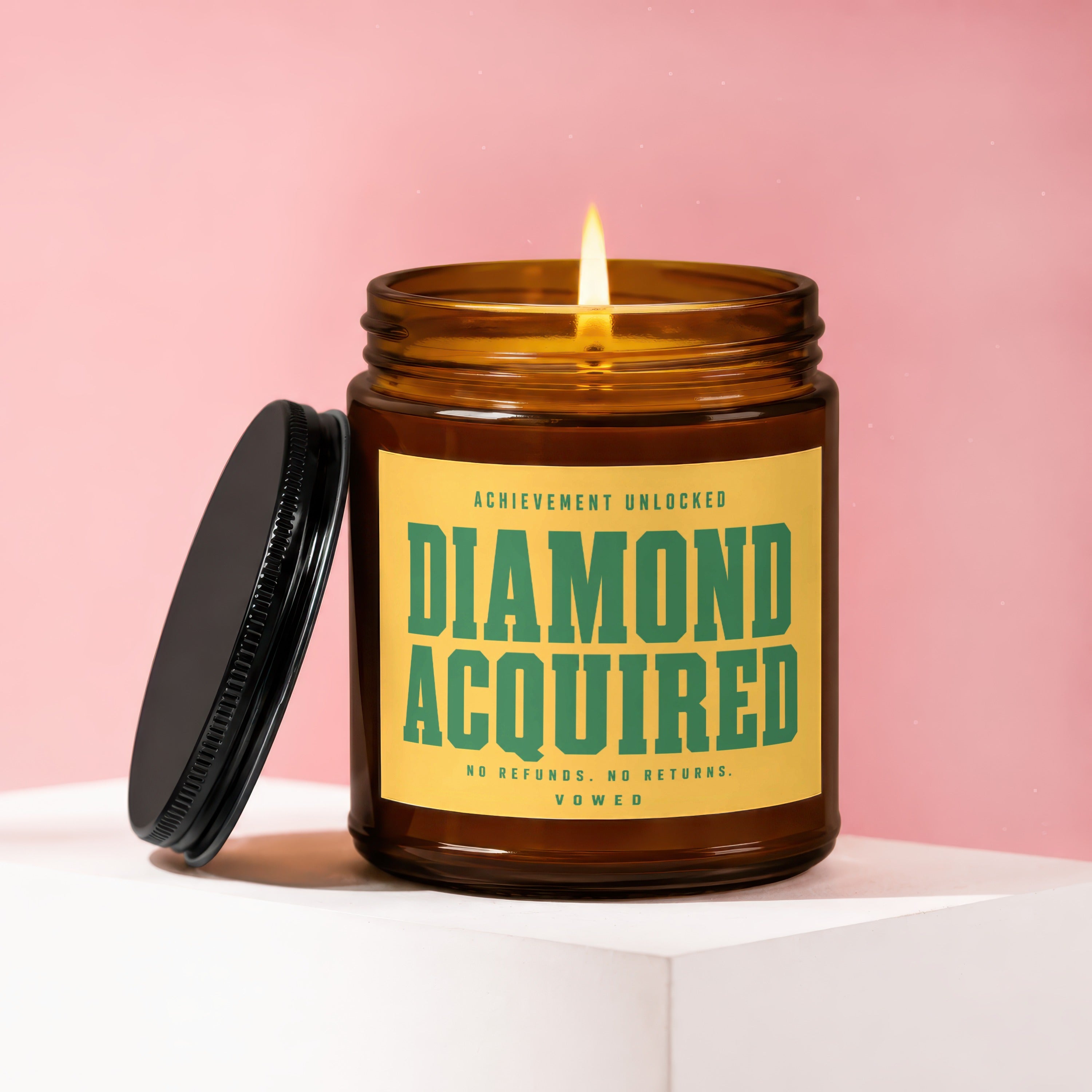 Lit amber glass candle with black lid, "DIAMOND ACQUIRED" text.