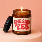 Burning candle in amber jar with "SHE SAID YES" and "VOWED" text on label.