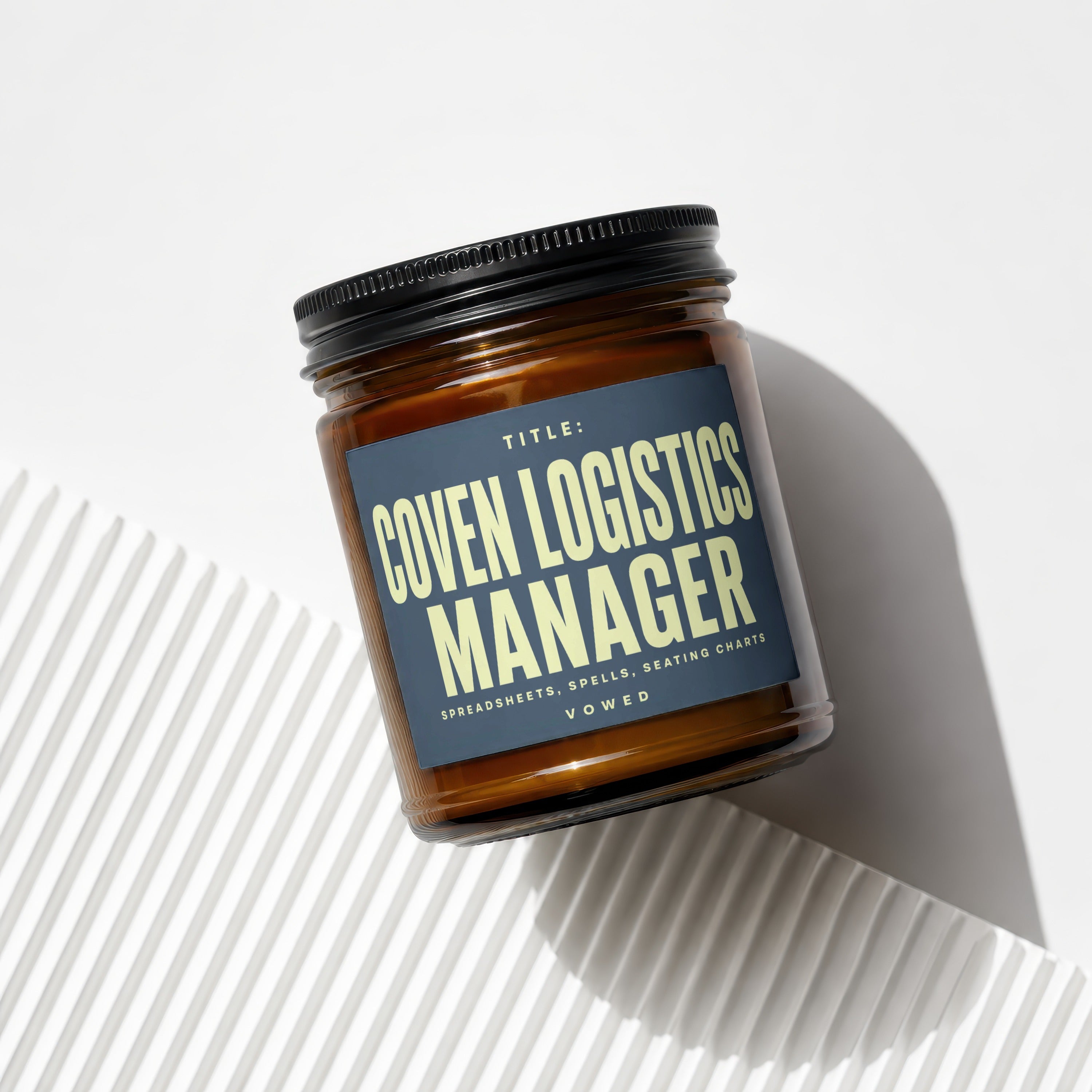 Amber jar candle with a dark blue label reading "TITLE: COVEN LOGISTICS MANAGER SPREADSHEETS, SPELLS, SEATING CHARTS VOWED".
