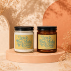 Two candles with "Love, But Make It Home" text on cream labels displayed on peach pedestals.
