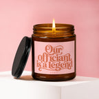 Amber candle with lit wick and pink label reading "Our officiant is a legend".