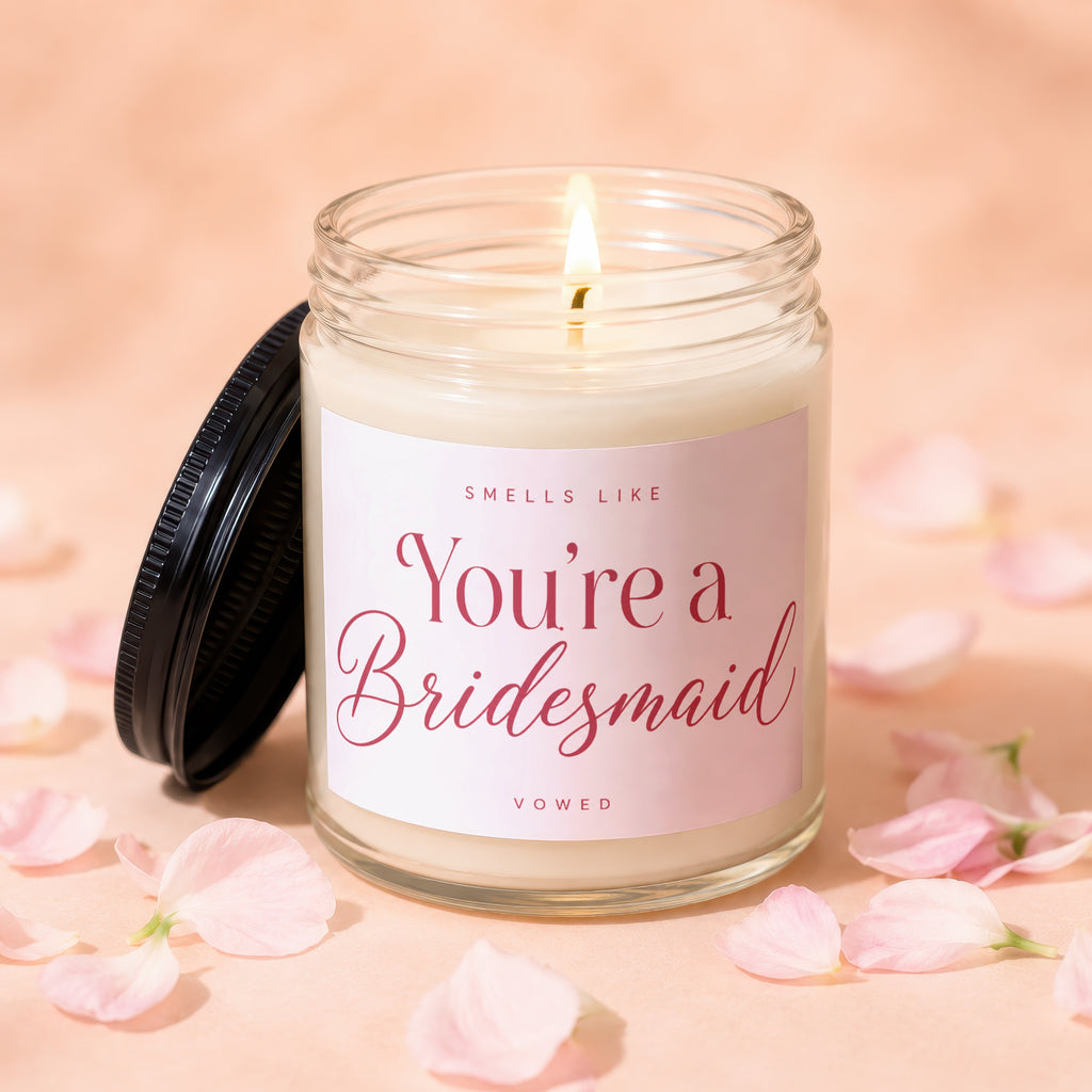 Glass candle with lit wick, pink label reads "Smells Like You're a Bridesmaid VOWED."