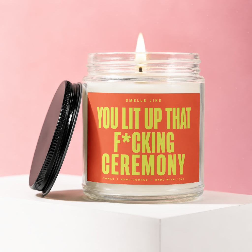 Glass candle with a black lid, lit wick, and orange label saying "You Lit Up That F*cking Ceremony".