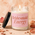 Lit scented candle in glass jar with pink label reading "Smells Like Bridesmaid Energy".