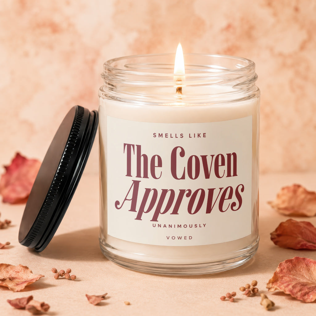 Glass jar candle with lit wick and label reading "SMELLS LIKE The Coven Approves UNANIMOUSLY VOWED".