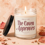 Glass jar candle with lit wick and label reading "SMELLS LIKE The Coven Approves UNANIMOUSLY VOWED".