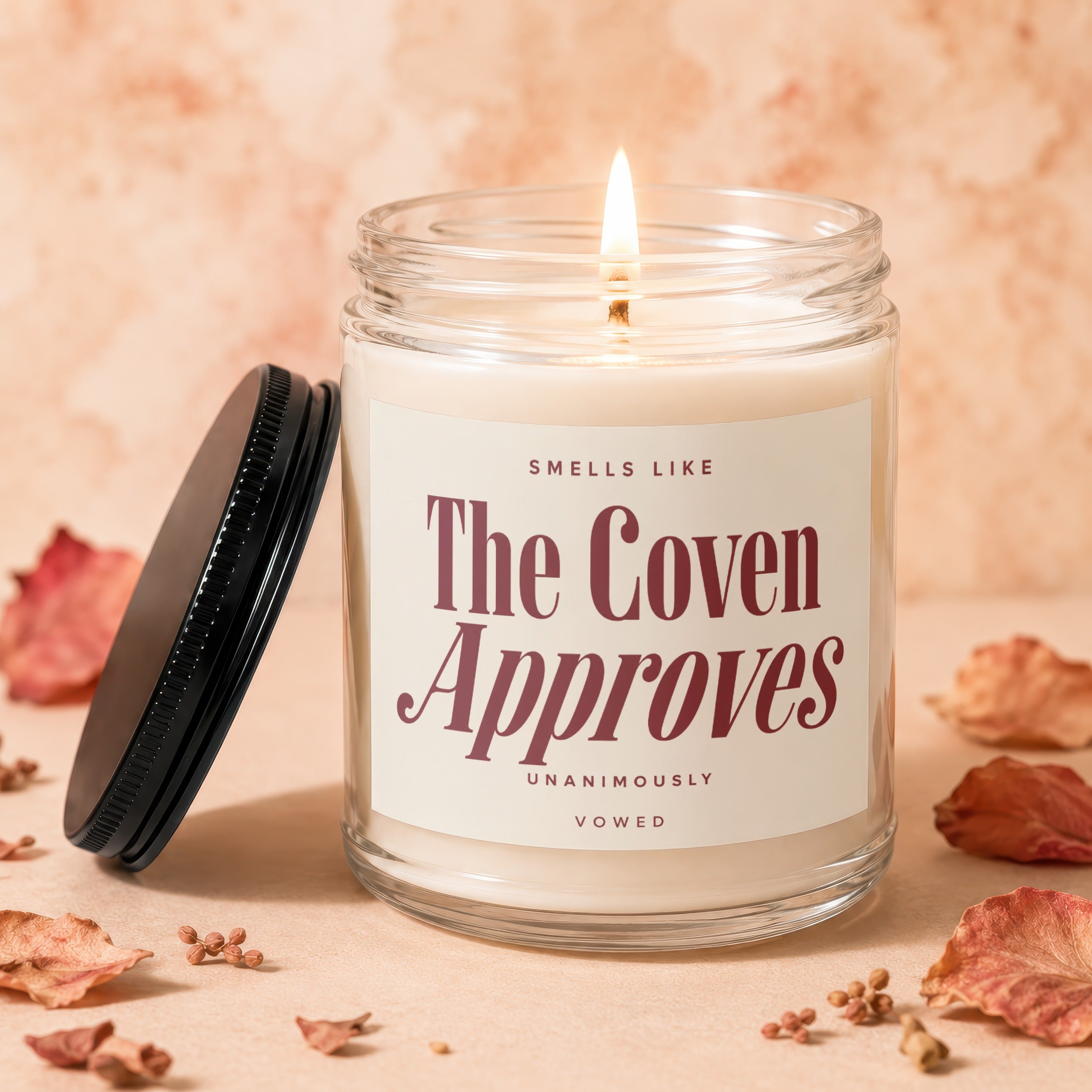 Glass jar candle with lit wick and label reading "SMELLS LIKE The Coven Approves UNANIMOUSLY VOWED".
