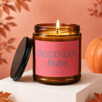 Amber candle with "GOOD LUCK, Babe." text on pink label, lit flame, black lid.