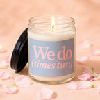 Jar candle with lit wick, pink text "We do (times two)" on label, and scattered petals.
