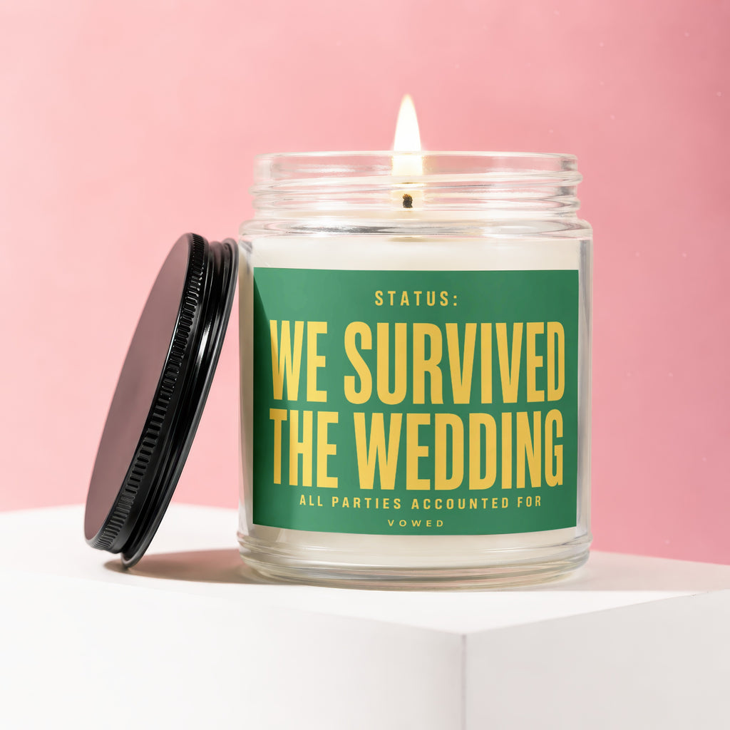 Glass candle with a lit wick, featuring a green label that reads "STATUS: WE SURVIVED THE WEDDING ALL PARTIES ACCOUNTED FOR VOWED".