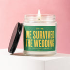 Glass candle with a lit wick, featuring a green label that reads "STATUS: WE SURVIVED THE WEDDING ALL PARTIES ACCOUNTED FOR VOWED".