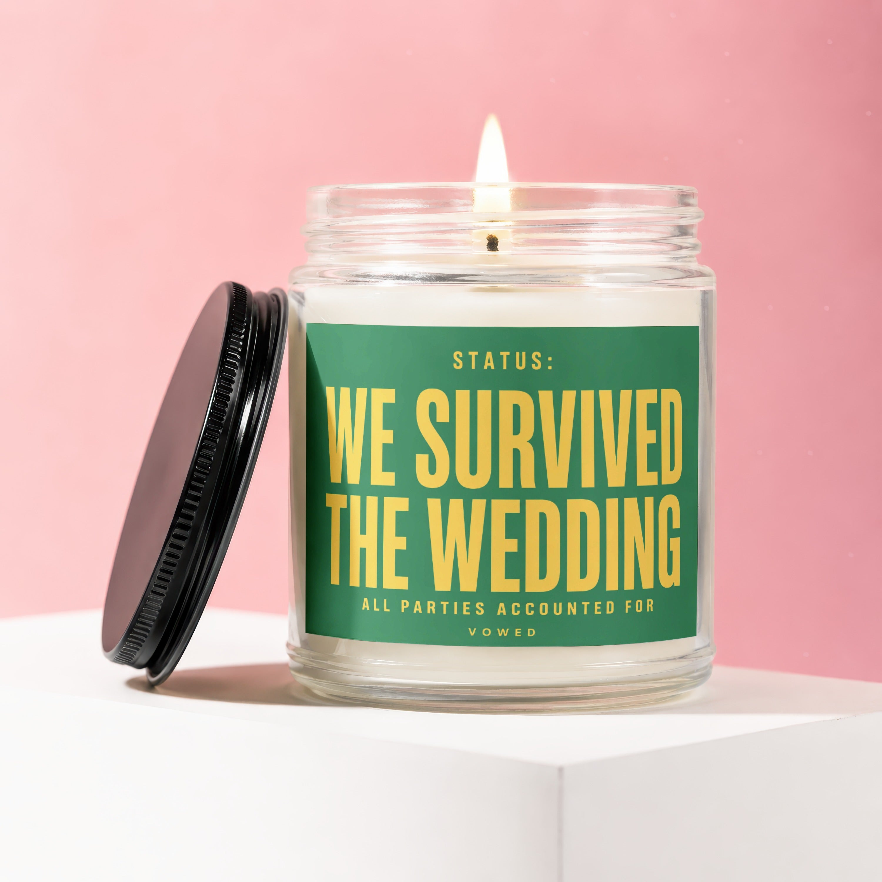 Glass candle with a lit wick, featuring a green label that reads "STATUS: WE SURVIVED THE WEDDING ALL PARTIES ACCOUNTED FOR VOWED".