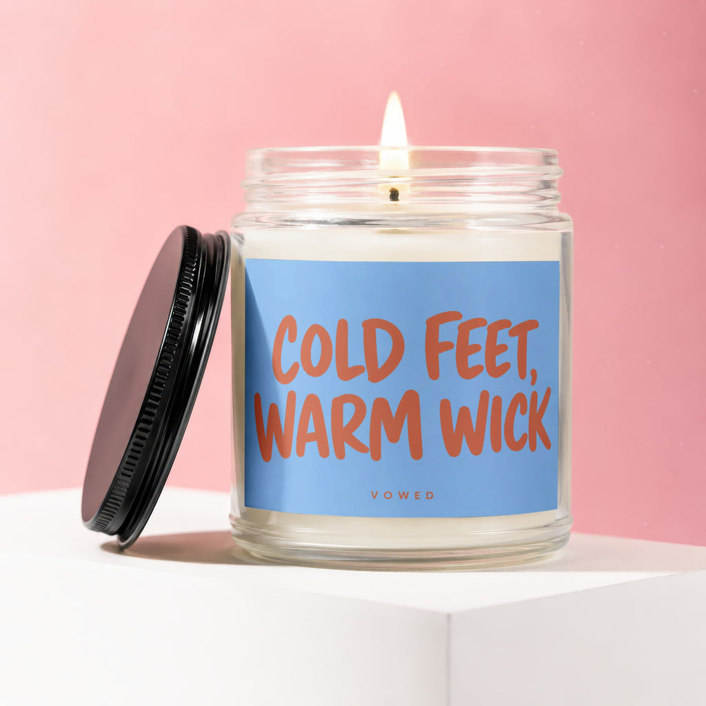 Glass jar candle with lit wick and lid, featuring "COLD FEET, WARM WICK" label.