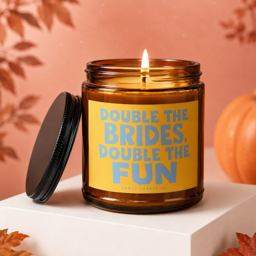 Burning amber glass candle with yellow label reading "DOUBLE THE BRIDES, DOUBLE THE FUN."