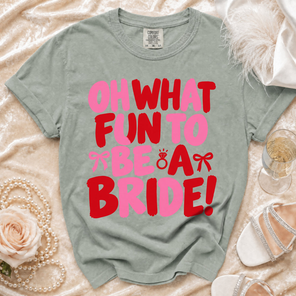 Sage green t-shirt printed with red and pink text reading Oh What Fun To Be A Bride surrounded by wedding accessories.