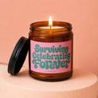 Amber jar candle with lit flame and pink label reading "Surviving Celebrating Forever Vowed".