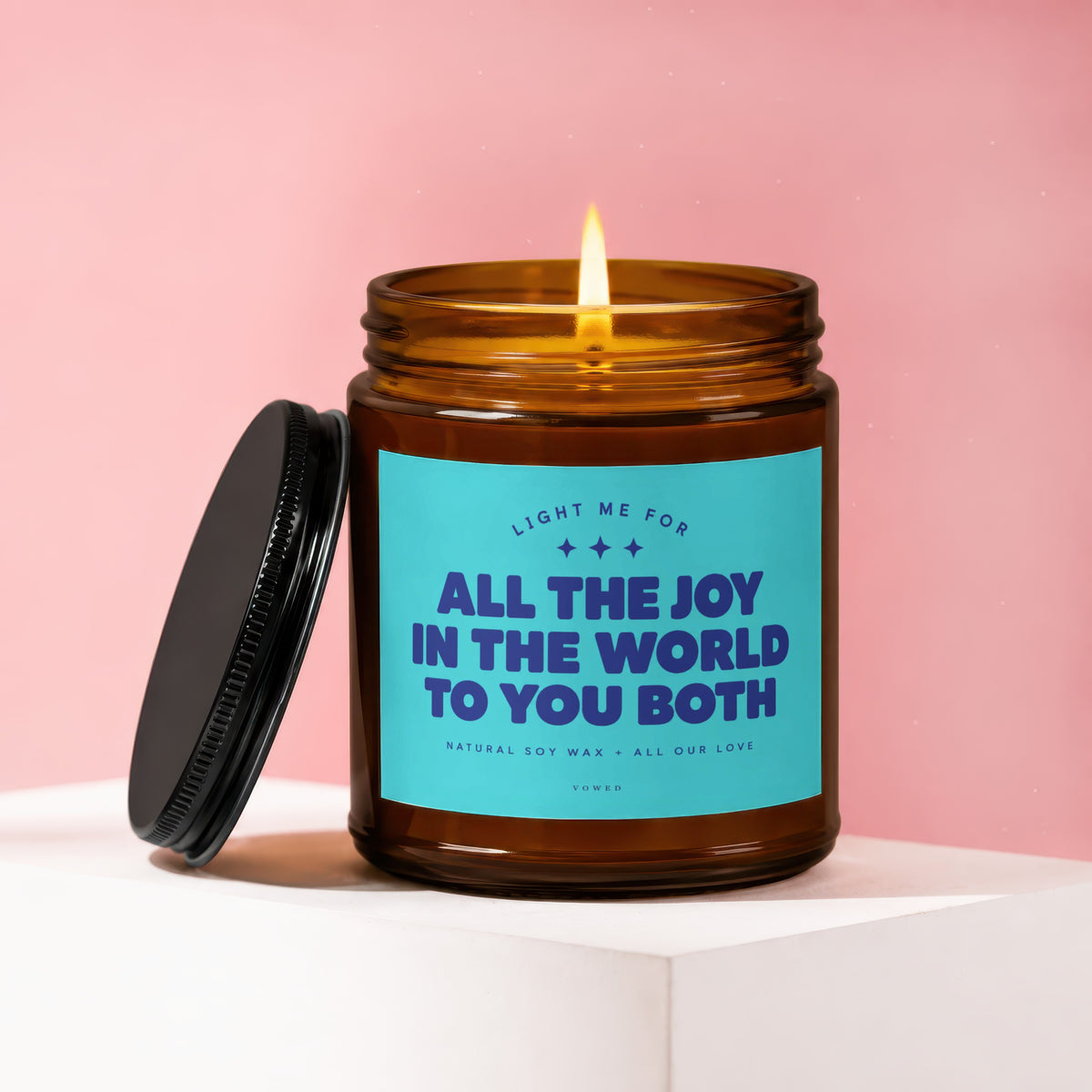 Lit amber glass candle with a teal label reading "ALL THE JOY IN THE WORLD TO YOU BOTH".