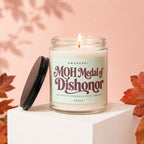 Glass candle labeled "MOH Medal of Dishonor" with lid off, lit wick.