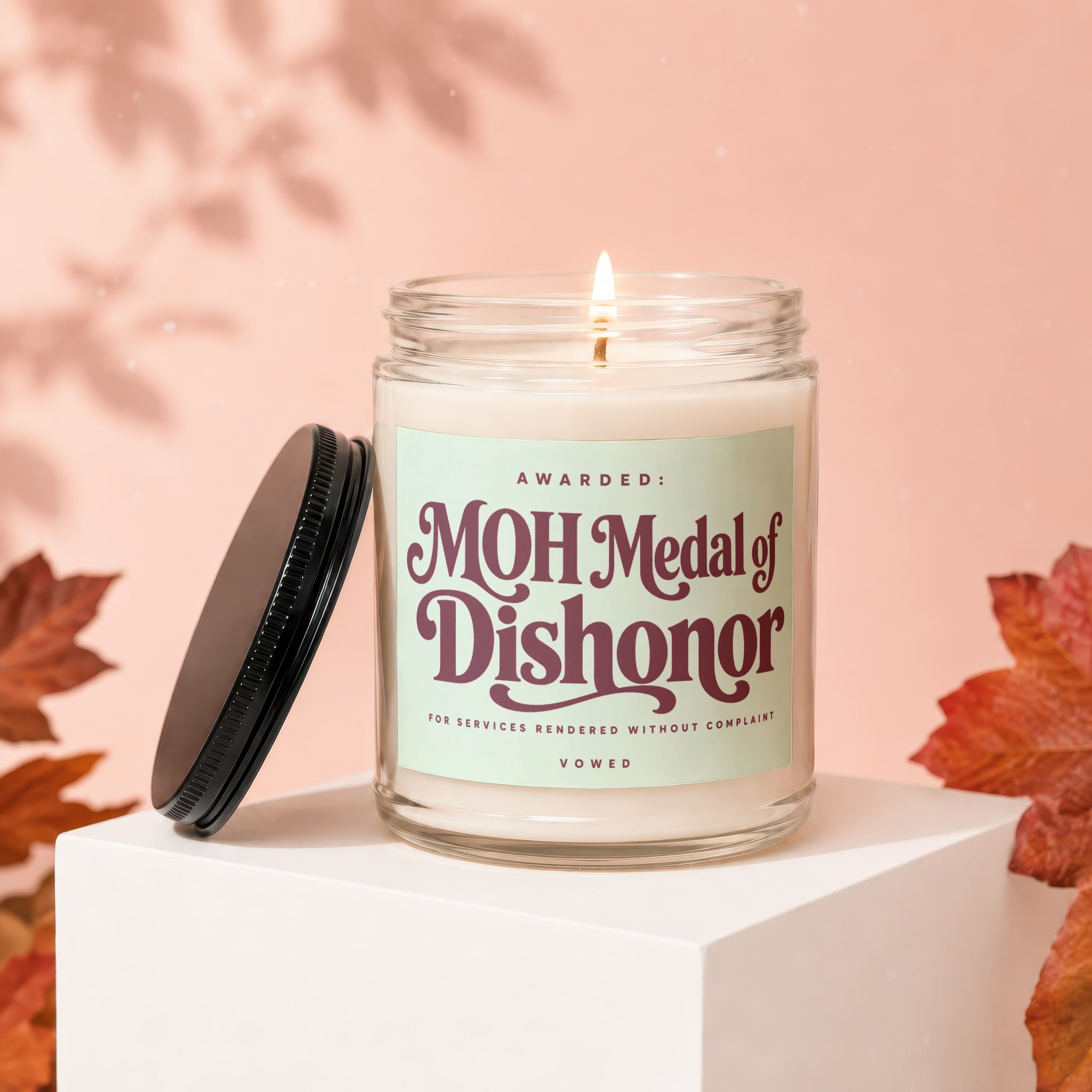 Glass candle labeled "MOH Medal of Dishonor" with lid off, lit wick.