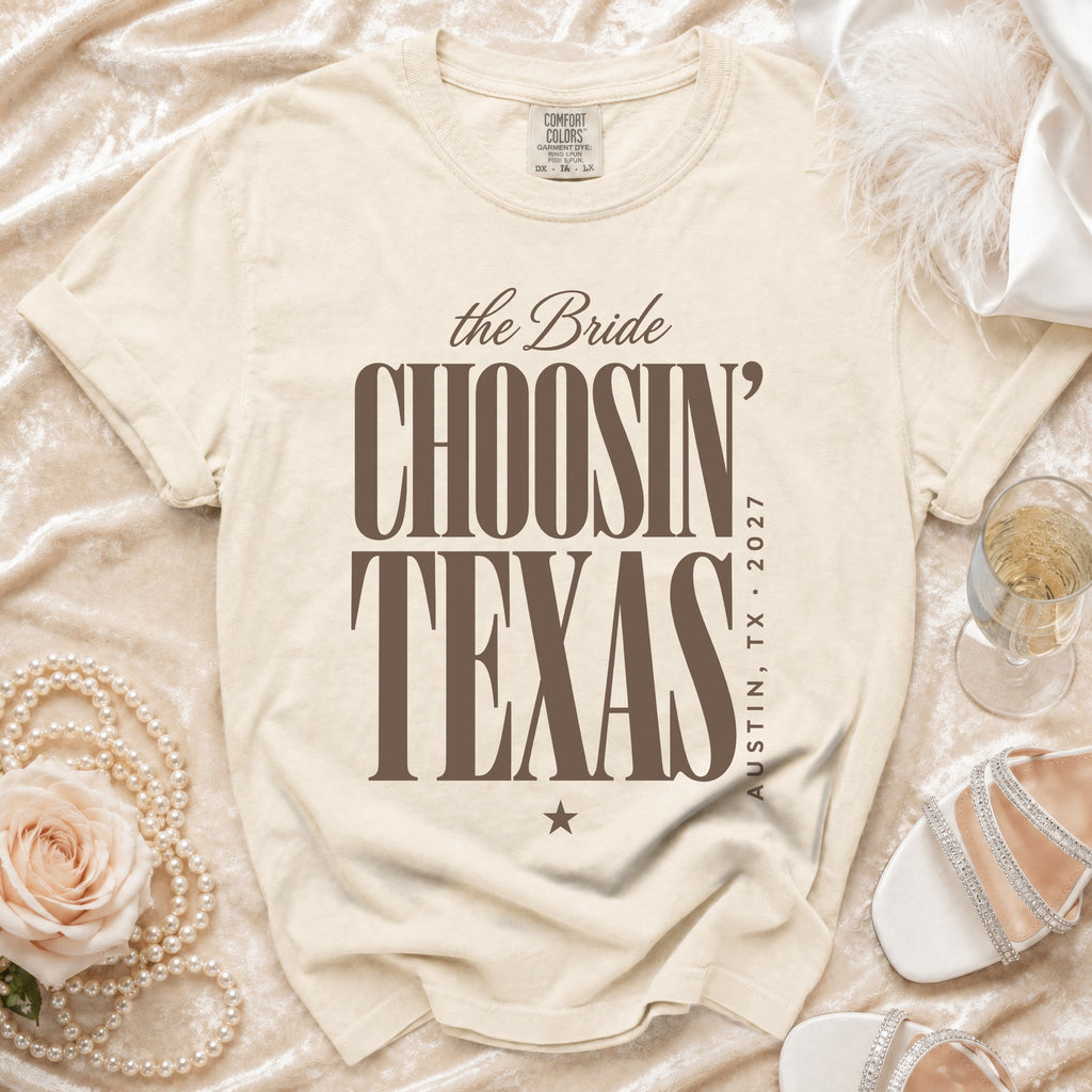 A cream-colored t-shirt with brown text reading "the Bride CHOOSIN' TEXAS AUSTIN, TX - 2027" lies on a velvet surface with accessories.