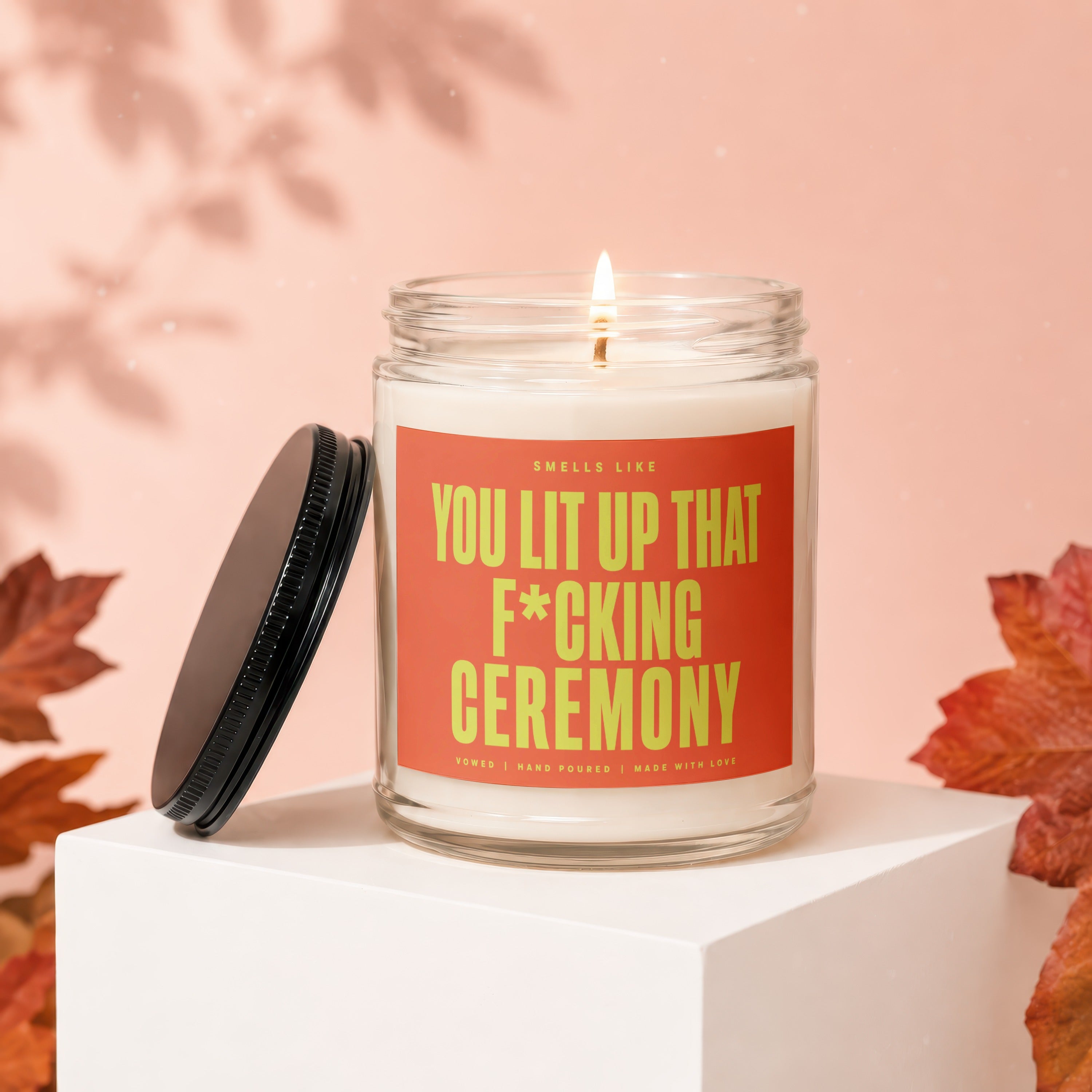 Glass candle with lit wick and orange label reading "Smells Like You Lit Up That F*cking Ceremony"