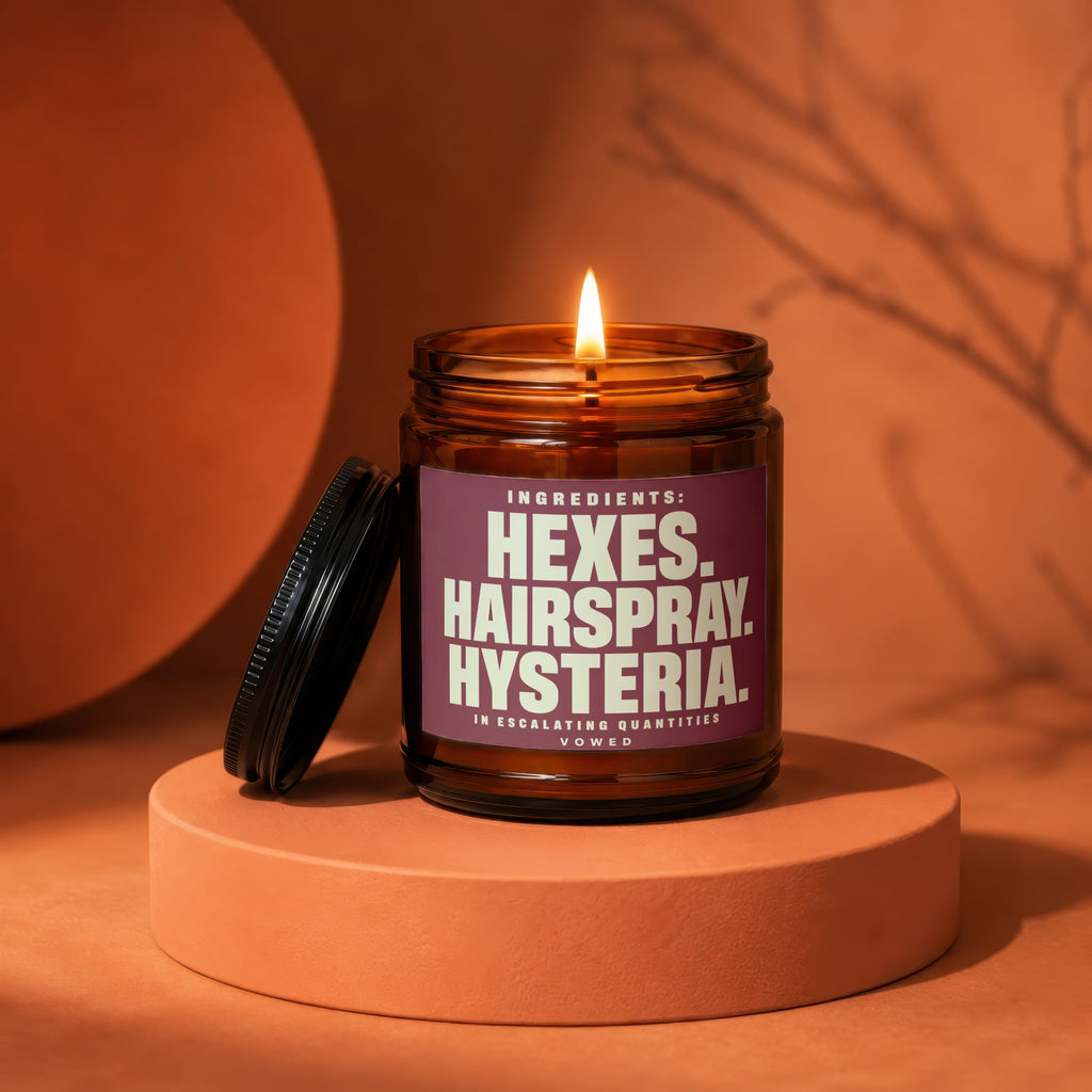 Amber glass candle with lit flame displays label with "Hexes. Hairspray. Hysteria."