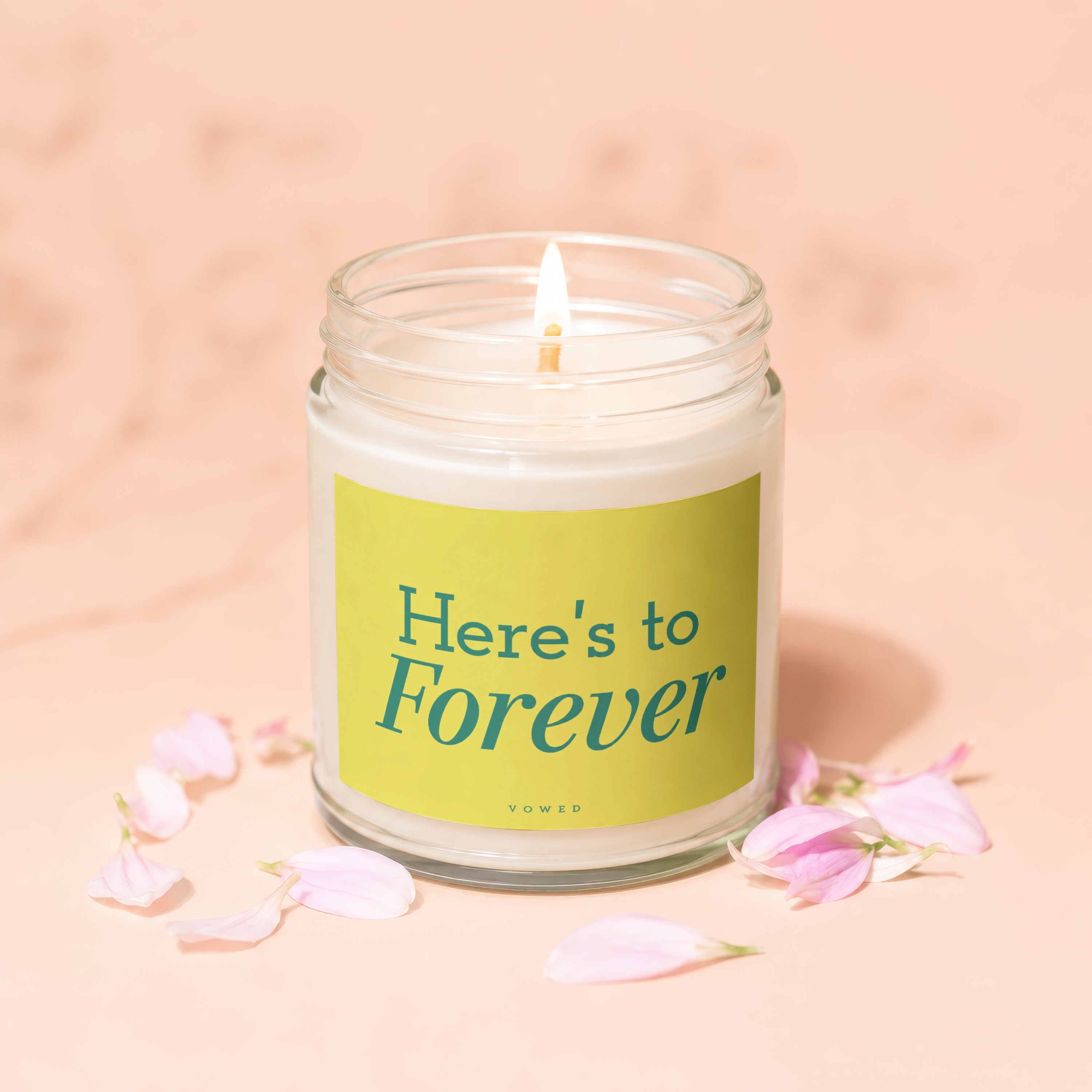 Jar candle with lit flame, green label reads "Here's to Forever", next to pink petals.