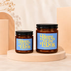 Two amber glass candles with blue labels that read "Here for the I Do's".