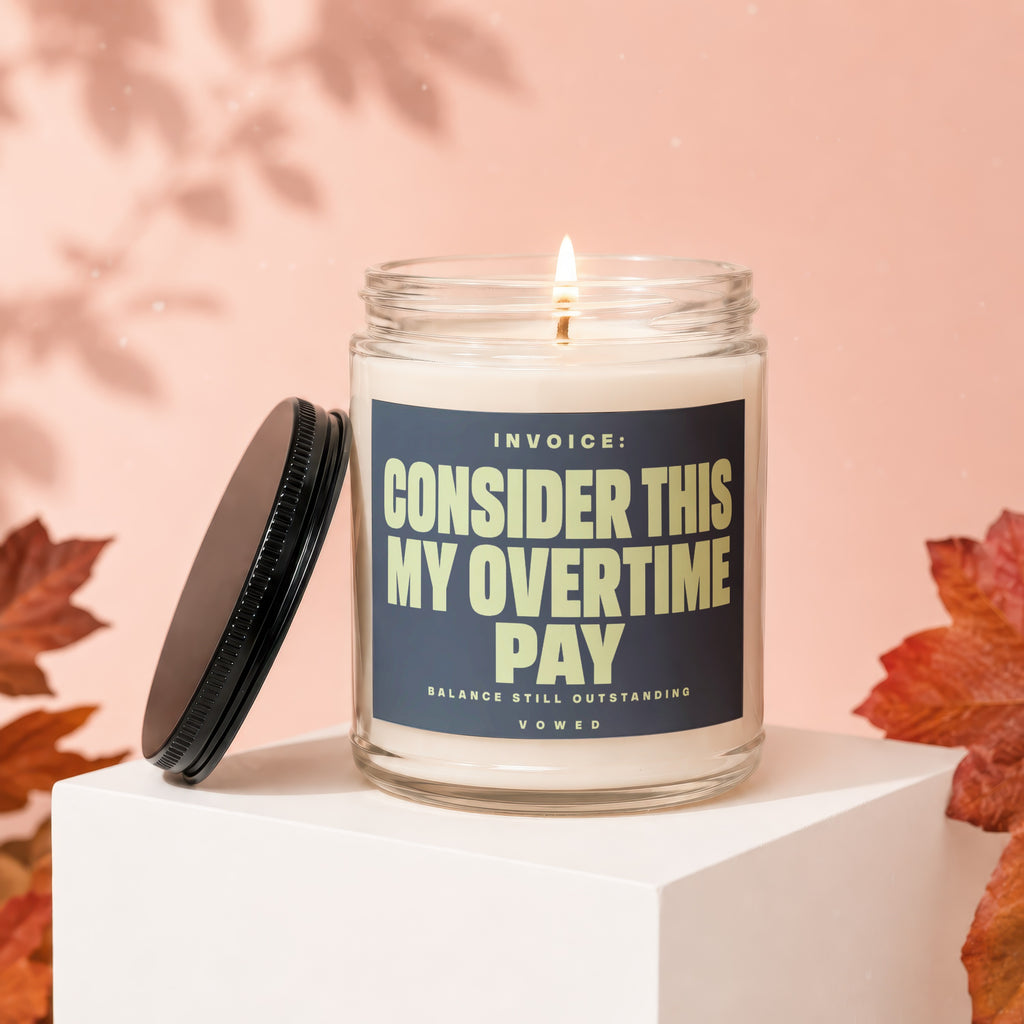 Glass candle with lit wick, lid off, featuring text "INVOICE: CONSIDER THIS MY OVERTIME PAY".