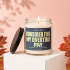 Glass candle with lit wick, lid off, featuring text "INVOICE: CONSIDER THIS MY OVERTIME PAY".