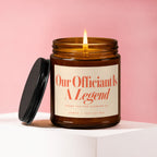 Amber jar candle with lit wick reads "Our Officiant Is A Legend".