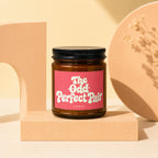 Brown glass candle with pink label reading "The Odd Perfect Pair"