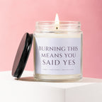 Candle with lit wick on white surface, black lid to the side, pink background.